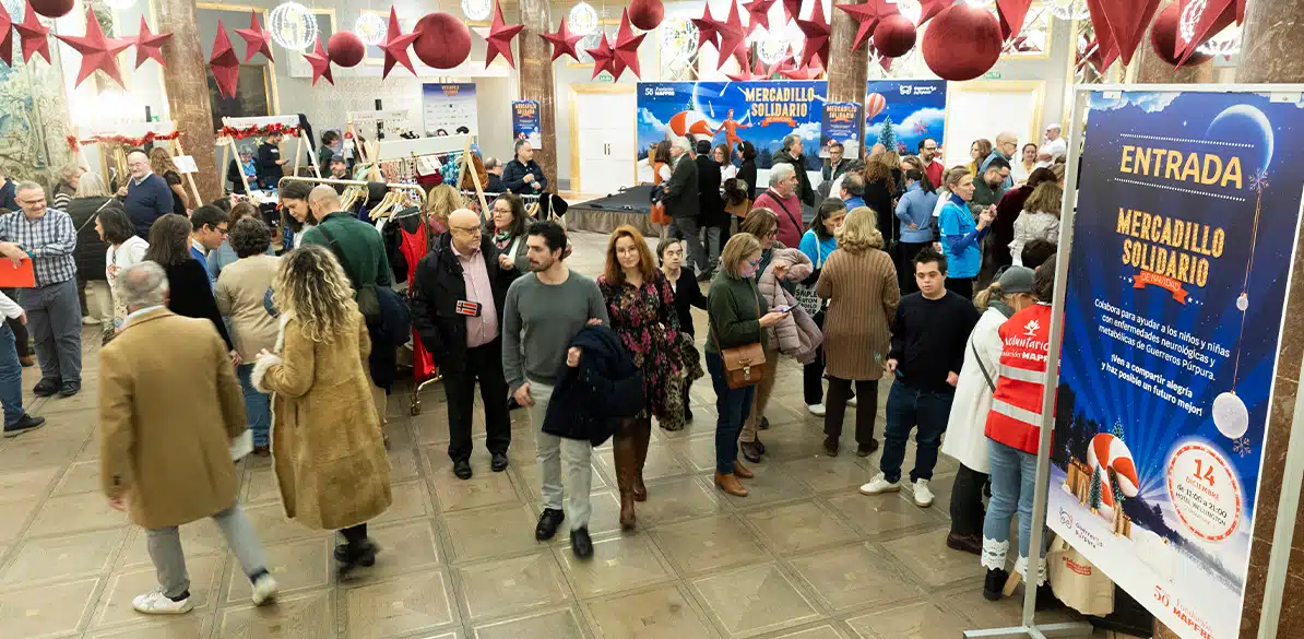 Mercadillo Solidario Vista general del Mercadillo Solidario con gran afluencia de personas, puestos decorados con motivos navideños y asistentes participando en la rifa solidaria, creando un ambiente festivo, solidario y de apoyo colectivo a una causa benéfica.