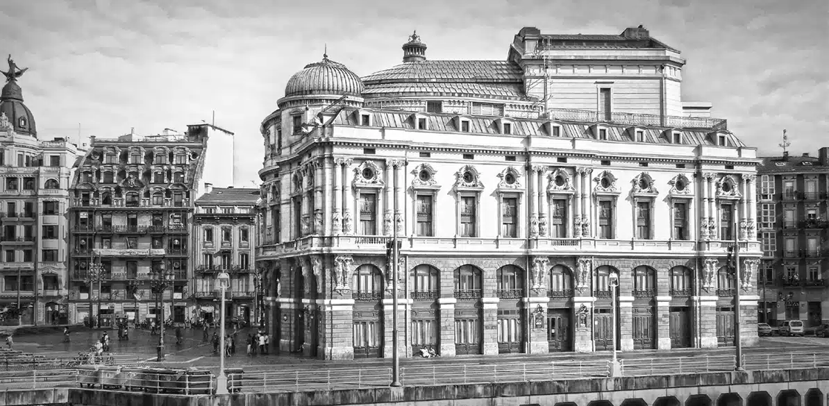 Largo e rubato Imagen en blanco y negro del Teatro Arriaga en Bilbao, un edificio neobarroco con cúpulas y fachada ornamentada. A su alrededor se ven edificios antiguos y personas caminando junto a la ría, creando una escena urbana histórica y viva.