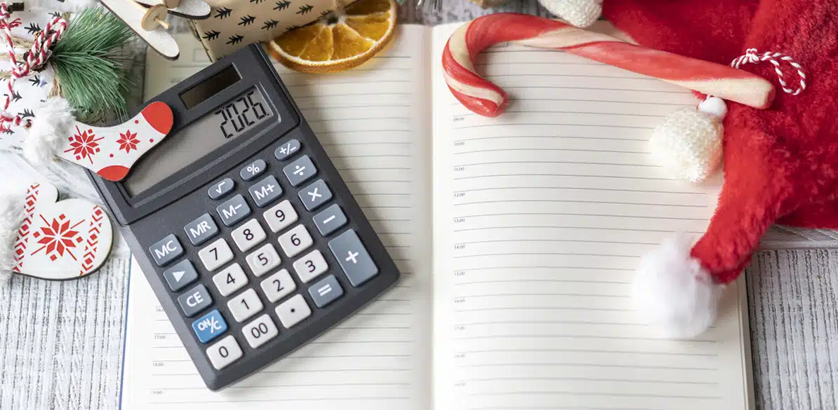 El mejor regalo para fin de año Calculadora gris encendida con el número 2026 sobre una agenda en blanco abierta, rodeada de regalos, rodajas de naranja seca, bastón de caramelo, guantes y gorro de Papá Noel, representando la planificación financiera y el control del gasto en las fiestas de fin de año.