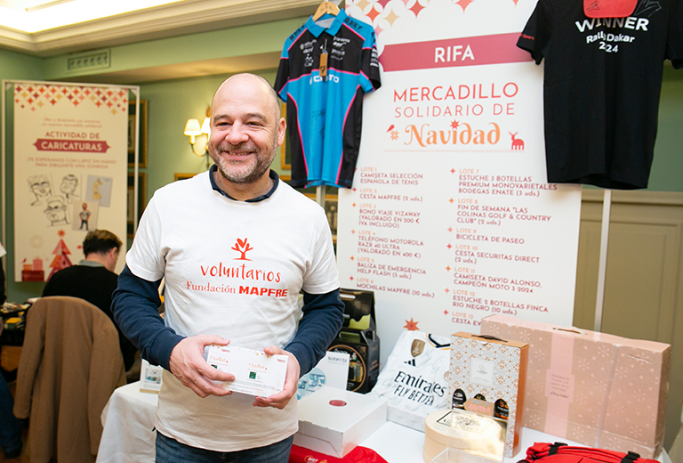 Rifa Solidaria Voluntario sonriente de Fundación MAPFRE sosteniendo boletos de una rifa durante el Mercadillo Solidario de Navidad, con mesas, premios y carteles informativos del evento al fondo.