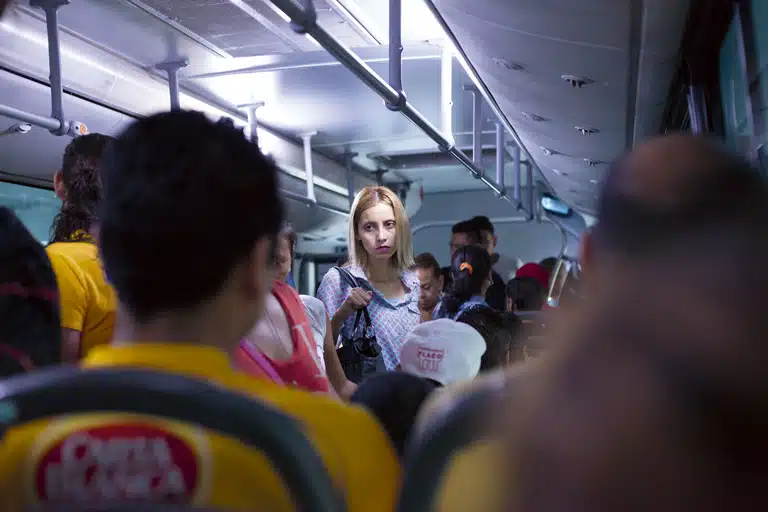 Alejandro Cartagena Interior de un autobús lleno de pasajeros, con una mujer rubia de pie en el centro mirando hacia la cámara entre asientos y personas desenfocadas.