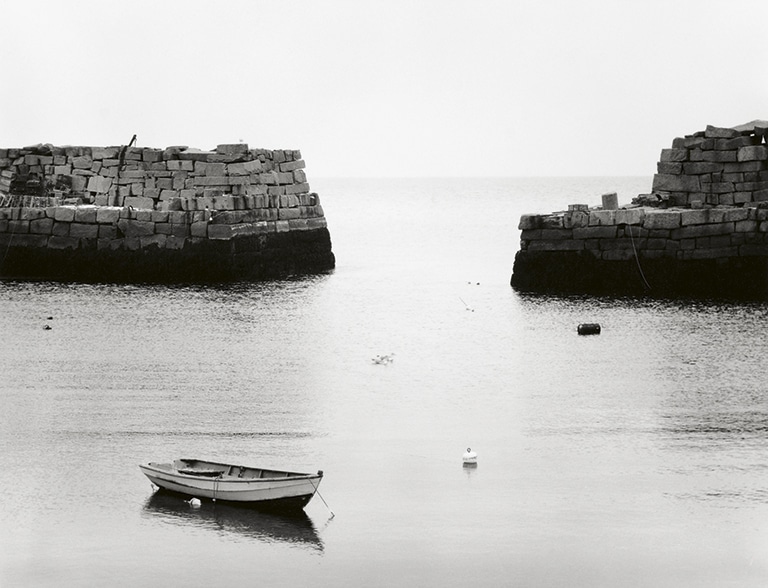 Minor White Barca sobre aguas tranquilas entre dos muelles de piedra en una fotografía en blanco y negro.