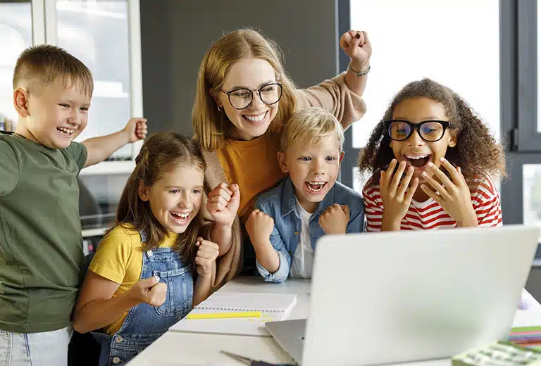 Materiales educativos Profesora y grupo de niños celebrando juntos mientras están frente a un ordenador portátil.